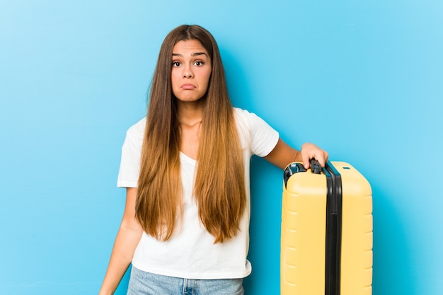 Joven mujer caucásica sosteniendo una maleta de viaje se encoge de hombros y abre los ojos confundidos.