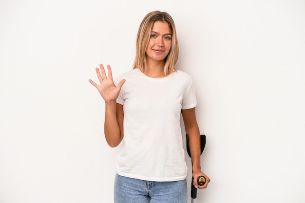 Joven mujer caucásica sosteniendo muleta aislado sobre fondo blanco sonriendo alegre mostrando el número cinco con los dedos.