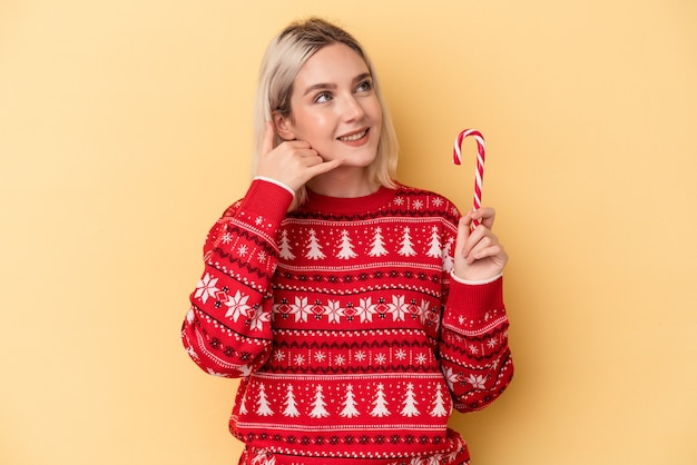 Joven mujer caucásica sosteniendo un palo de Navidad aislado sobre fondo amarillo que muestra un gesto de llamada de teléfono móvil con los dedos.