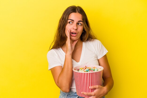 Joven mujer caucásica sosteniendo palomitas de maíz aislado sobre fondo amarillo está diciendo una noticia secreta de frenado caliente y mirando a un lado