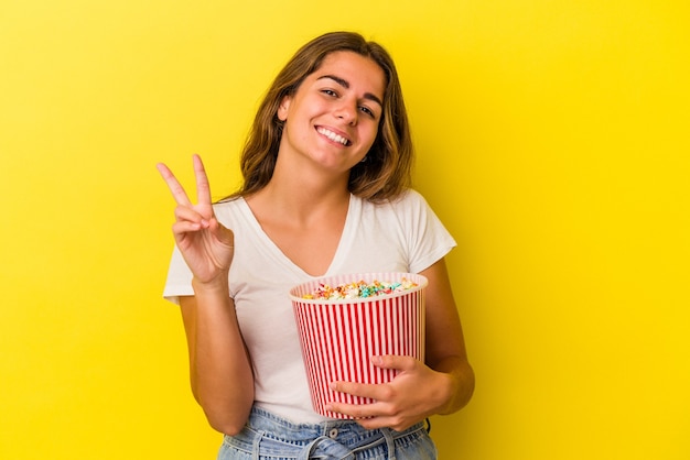 Joven mujer caucásica sosteniendo palomitas de maíz aislado sobre fondo amarillo que muestra el número dos con los dedos.