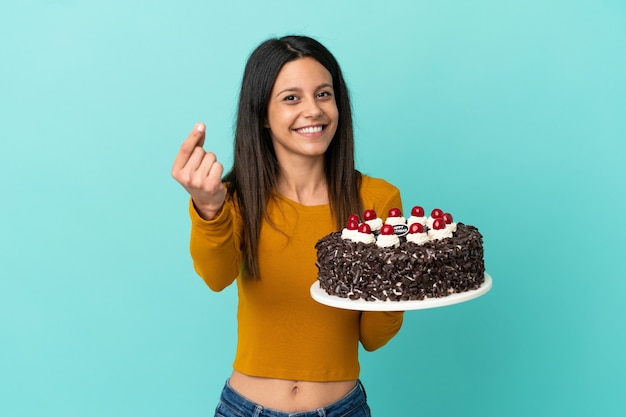 Joven mujer caucásica sosteniendo pastel de cumpleaños aislado sobre fondo azul haciendo gesto de dinero