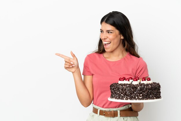 Joven mujer caucásica sosteniendo pastel de cumpleaños aislado sobre fondo blanco apuntando con el dedo hacia el lado y presentando un producto