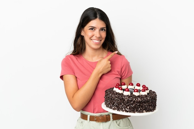 Joven mujer caucásica sosteniendo pastel de cumpleaños aislado sobre fondo blanco apuntando hacia el lado para presentar un producto