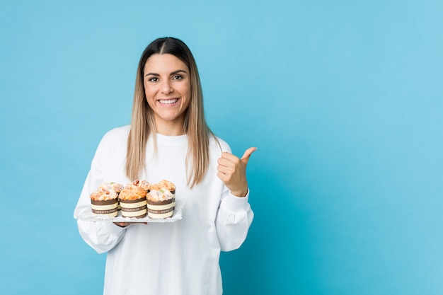 Joven mujer caucásica sosteniendo un pastel de dulces sonriendo y levantando el pulgar hacia arriba