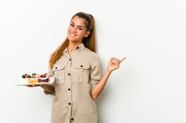 Joven mujer caucásica sosteniendo pasteles dulces sonriendo y apuntando a un lado, mostrando algo en el espacio en blanco.