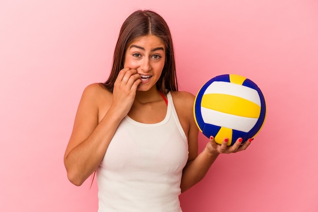 Joven mujer caucásica sosteniendo una pelota de voleibol aislada en la pared rosa mordiéndose las uñas, nerviosa y muy ansiosa.