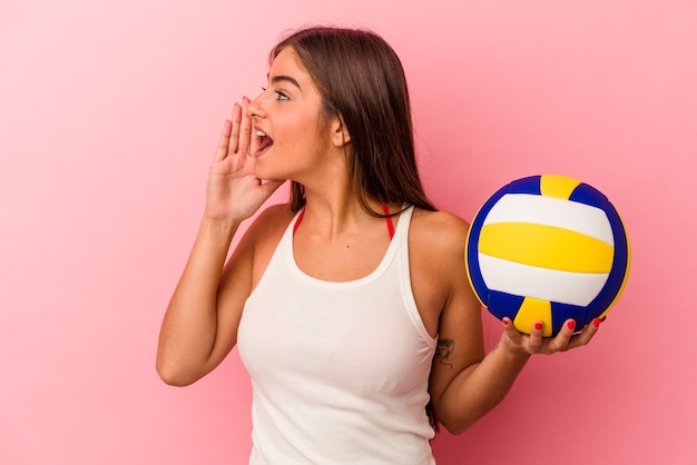 Joven mujer caucásica sosteniendo una pelota de voleibol aislada sobre fondo rosa gritando y sosteniendo la palma cerca de la boca abierta.