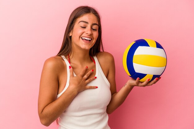 Joven mujer caucásica sosteniendo una pelota de voleibol aislada sobre fondo rosa se ríe a carcajadas manteniendo la mano en el pecho.