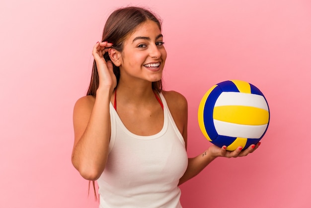 Joven mujer caucásica sosteniendo una pelota de voleibol aislada sobre fondo rosa tratando de escuchar un chisme.