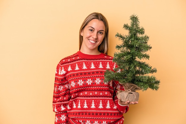 Joven mujer caucásica sosteniendo un pequeño árbol de Navidad aislado sobre fondo amarillo feliz, sonriente y alegre.