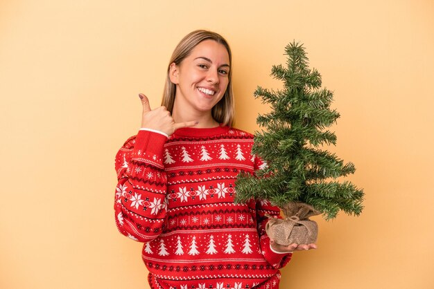 Joven mujer caucásica sosteniendo un pequeño árbol de Navidad aislado sobre fondo amarillo mostrando un gesto de llamada de teléfono móvil con los dedos.