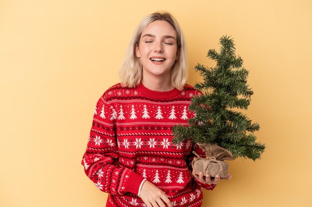 Joven mujer caucásica sosteniendo un pequeño árbol de Navidad aislado sobre fondo amarillo riendo y divirtiéndose.