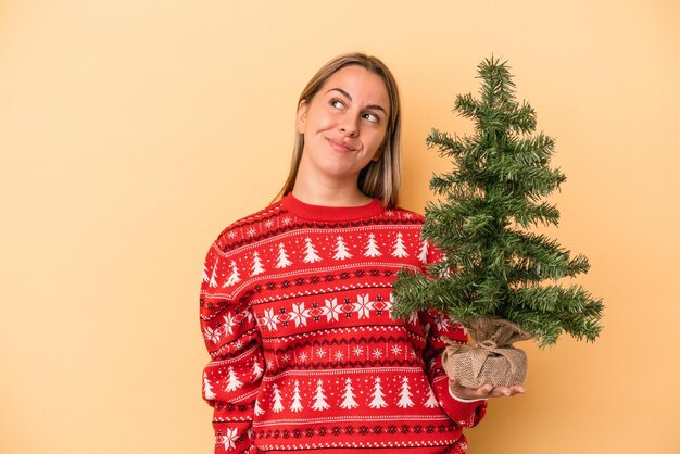 Joven mujer caucásica sosteniendo un pequeño árbol de Navidad aislado sobre fondo amarillo soñando con lograr metas y propósitos
