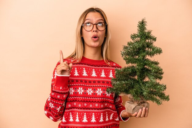 Joven mujer caucásica sosteniendo un pequeño árbol de Navidad aislado sobre fondo beige apuntando al revés con la boca abierta.