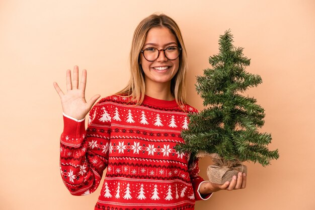 Joven mujer caucásica sosteniendo un pequeño árbol de Navidad aislado sobre fondo beige sonriendo alegre mostrando el número cinco con los dedos.