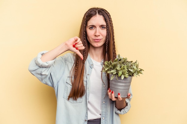 Joven mujer caucásica sosteniendo una planta aislada sobre fondo amarillo mostrando un gesto de aversión, pulgares hacia abajo. Concepto de desacuerdo.