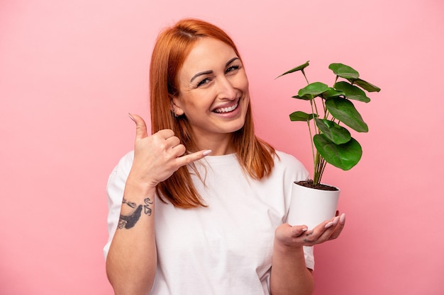 Joven mujer caucásica sosteniendo una planta aislada sobre fondo rosa Joven mujer caucásica sosteniendo una planta aislada sobre fondo rosa mostrando un gesto de llamada de teléfono móvil con los dedos.