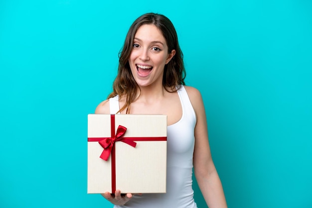 Joven mujer caucásica sosteniendo un regalo aislado de fondo azul con sorpresa y expresión facial conmocionada