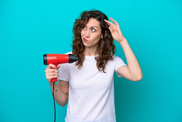 Joven mujer caucásica sosteniendo un secador de pelo aislado sobre fondo azul con dudas y con expresión de la cara confusa