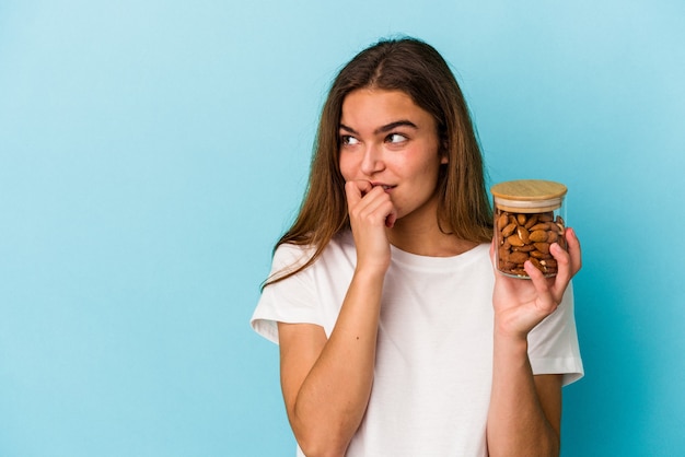 Joven mujer caucásica sosteniendo un tarro de almendras aislado sobre fondo azul relajado pensando en algo mirando un espacio de copia.