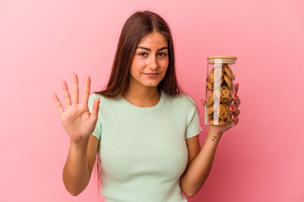 Joven mujer caucásica sosteniendo un tarro de galletas aislado sobre fondo rosa sonriendo alegre mostrando el número cinco con los dedos.