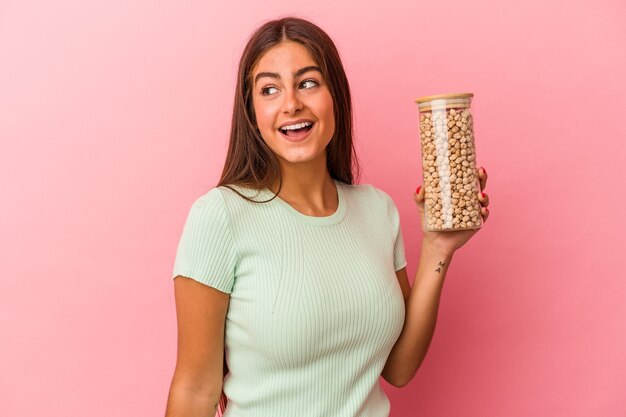 Joven mujer caucásica sosteniendo un tarro de garbanzos aislado sobre fondo rosa se ve a un lado sonriente, alegre y agradable.