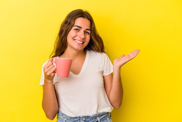 Joven mujer caucásica sosteniendo una taza aislada sobre fondo amarillo que muestra un espacio de copia en una palma y sosteniendo otra mano en la cintura.