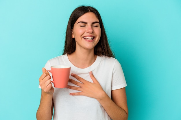 Joven mujer caucásica sosteniendo una taza aislada sobre fondo azul se ríe a carcajadas manteniendo la mano en el pecho.