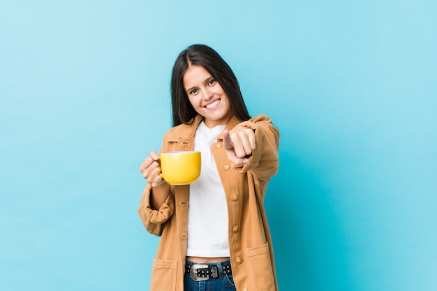 Joven mujer caucásica sosteniendo una taza de café con una sonrisa alegre apuntando al frente.