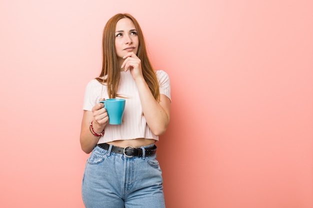 Joven mujer caucásica sosteniendo una taza mirando hacia los lados con expresión dudosa y escéptica.