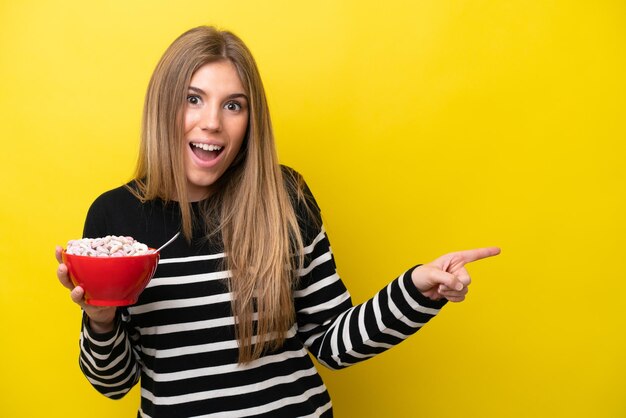 Joven mujer caucásica sosteniendo un tazón de cereales aislado en un fondo amarillo sorprendida y señalando con el dedo hacia un lado