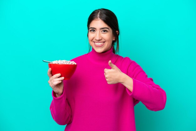 Joven mujer caucásica sosteniendo un tazón de cereales aislado de fondo azul con los pulgares hacia arriba porque algo bueno ha sucedido