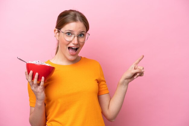 Joven mujer caucásica sosteniendo un tazón de cereales aislado en un fondo rosado sorprendida y señalando con el dedo hacia un lado