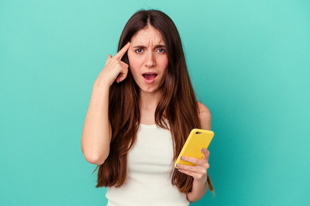 Joven mujer caucásica sosteniendo un teléfono móvil aislado sobre fondo azul mostrando un gesto de decepción con el dedo índice.