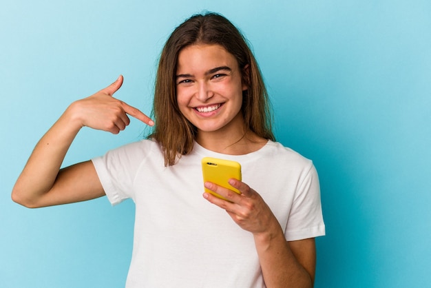 Joven mujer caucásica sosteniendo un teléfono móvil aislado sobre fondo azul persona apuntando con la mano a un espacio de copia de camisa, orgulloso y seguro