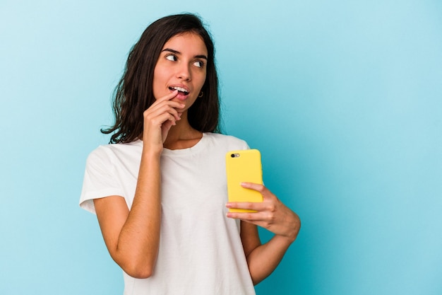 Joven mujer caucásica sosteniendo un teléfono móvil aislado sobre fondo azul relajado pensando en algo mirando un espacio de copia.