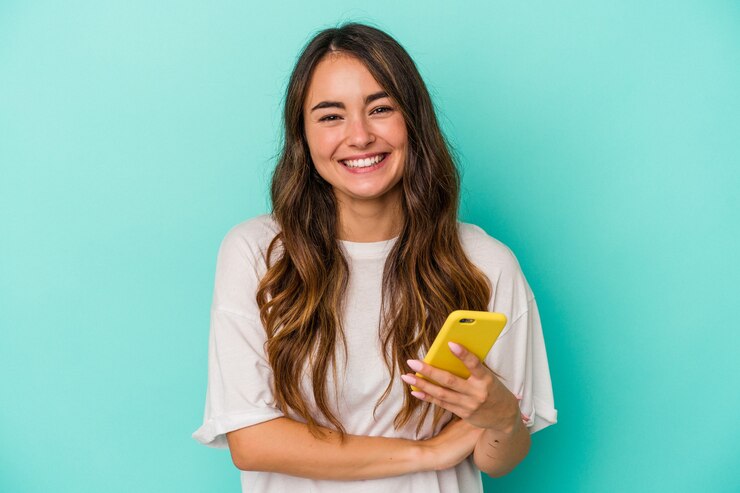 Foto joven mujer caucásica sosteniendo un teléfono móvil aislado sobre fondo azul riendo y divirtiéndose.