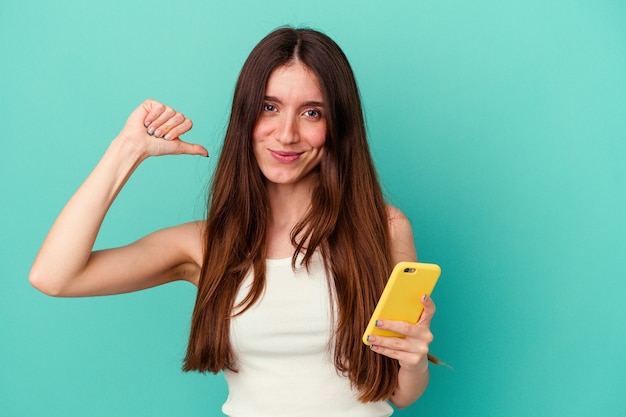 Joven mujer caucásica sosteniendo un teléfono móvil aislado sobre fondo azul se siente orgullosa y segura de sí misma, ejemplo a seguir.