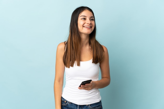 Joven mujer caucásica mediante teléfono móvil aislado en azul riendo