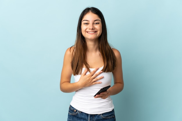 Joven mujer caucásica con teléfono móvil aislado en azul sonriendo mucho