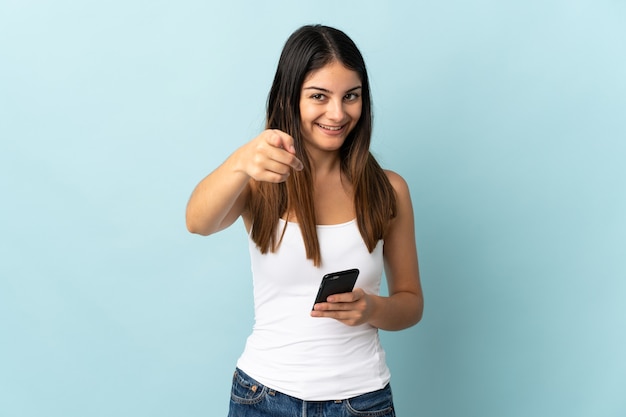 Joven mujer caucásica con teléfono móvil aislado en la pared azul sorprendido y apuntando hacia el frente