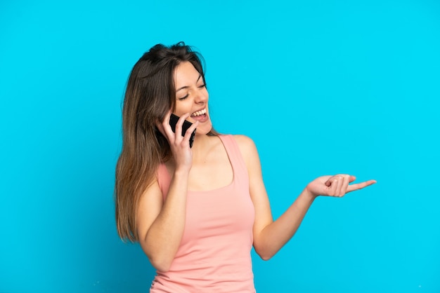 Joven mujer caucásica con teléfono móvil aislado sobre fondo azul apuntando con el dedo hacia el lado y presentando un producto