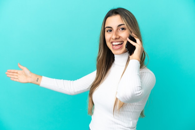 Foto joven mujer caucásica mediante teléfono móvil aislado sobre fondo azul extendiendo las manos hacia el lado para invitar a venir