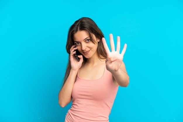 Joven mujer caucásica con teléfono móvil aislado sobre fondo azul feliz y contando cuatro con los dedos
