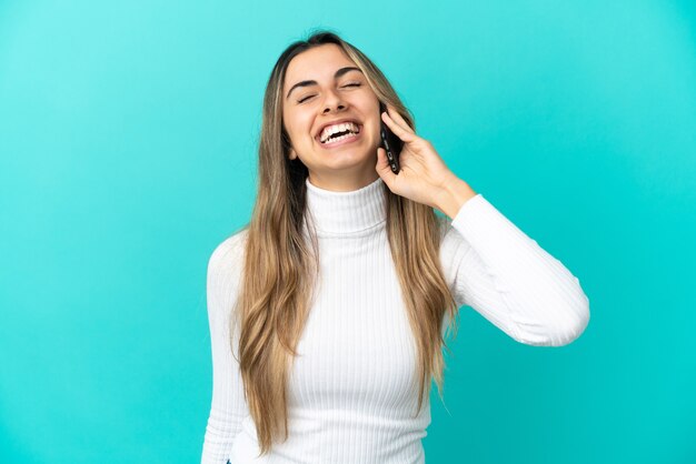 Joven mujer caucásica mediante teléfono móvil aislado sobre fondo azul riendo