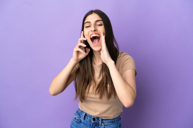 Joven mujer caucásica con teléfono móvil aislado sobre fondo púrpura gritando con la boca abierta