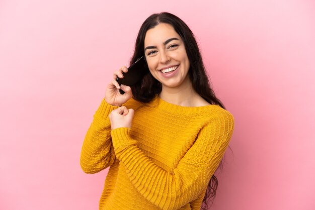 Joven mujer caucásica con teléfono móvil aislado sobre fondo rosa celebrando una victoria