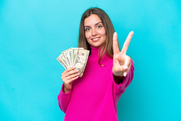 Joven mujer caucásica tomando mucho dinero aislada de fondo azul sonriendo y mostrando el signo de la victoria