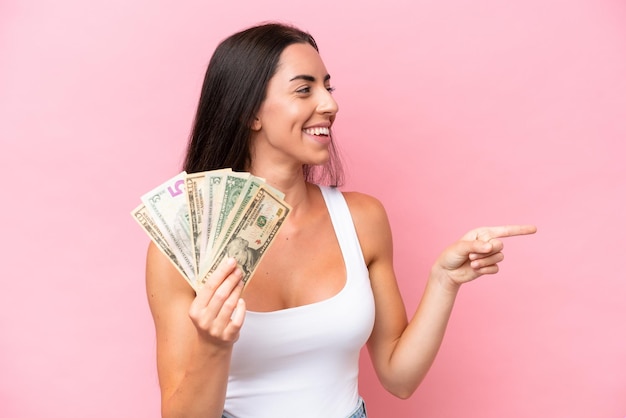 Joven mujer caucásica tomando mucho dinero aislada de fondo rosa apuntando hacia un lado para presentar un producto